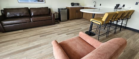 A modern living room with a brown leather sofa, a pink armchair, a wooden floor, a kitchen area with a sink and bar stools, and a framed picture on the wall.
