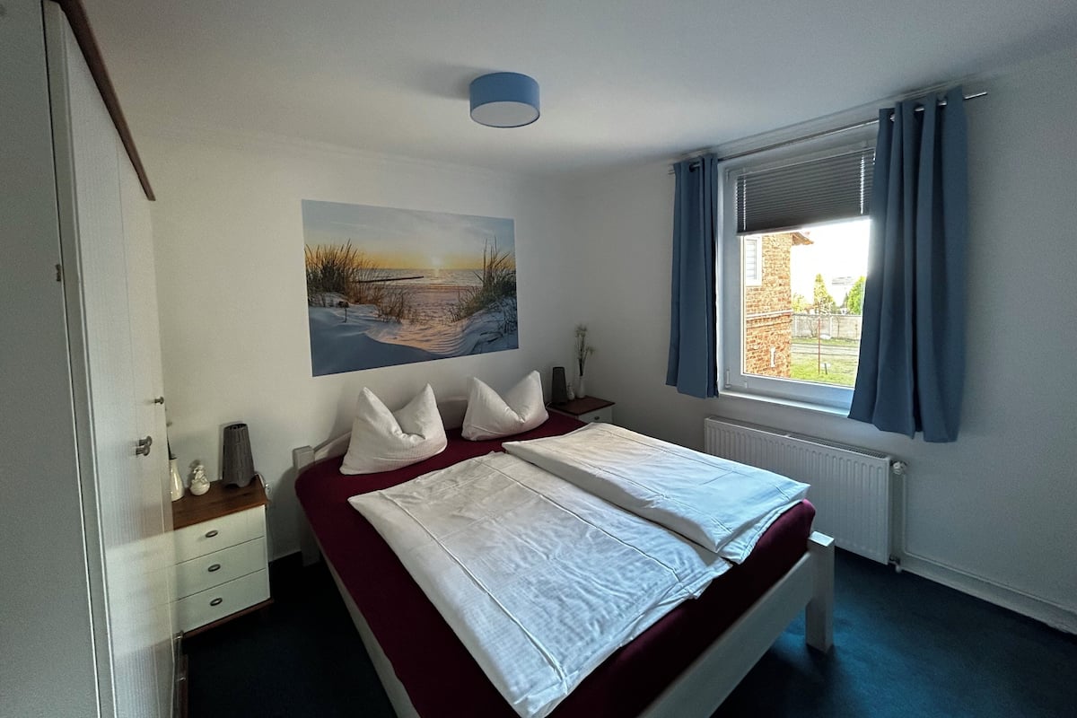 Une chambre à coucher comprenant un lit, une table de chevet, une lampe, un tableau, une fenêtre avec des rideaux et un radiateur.
