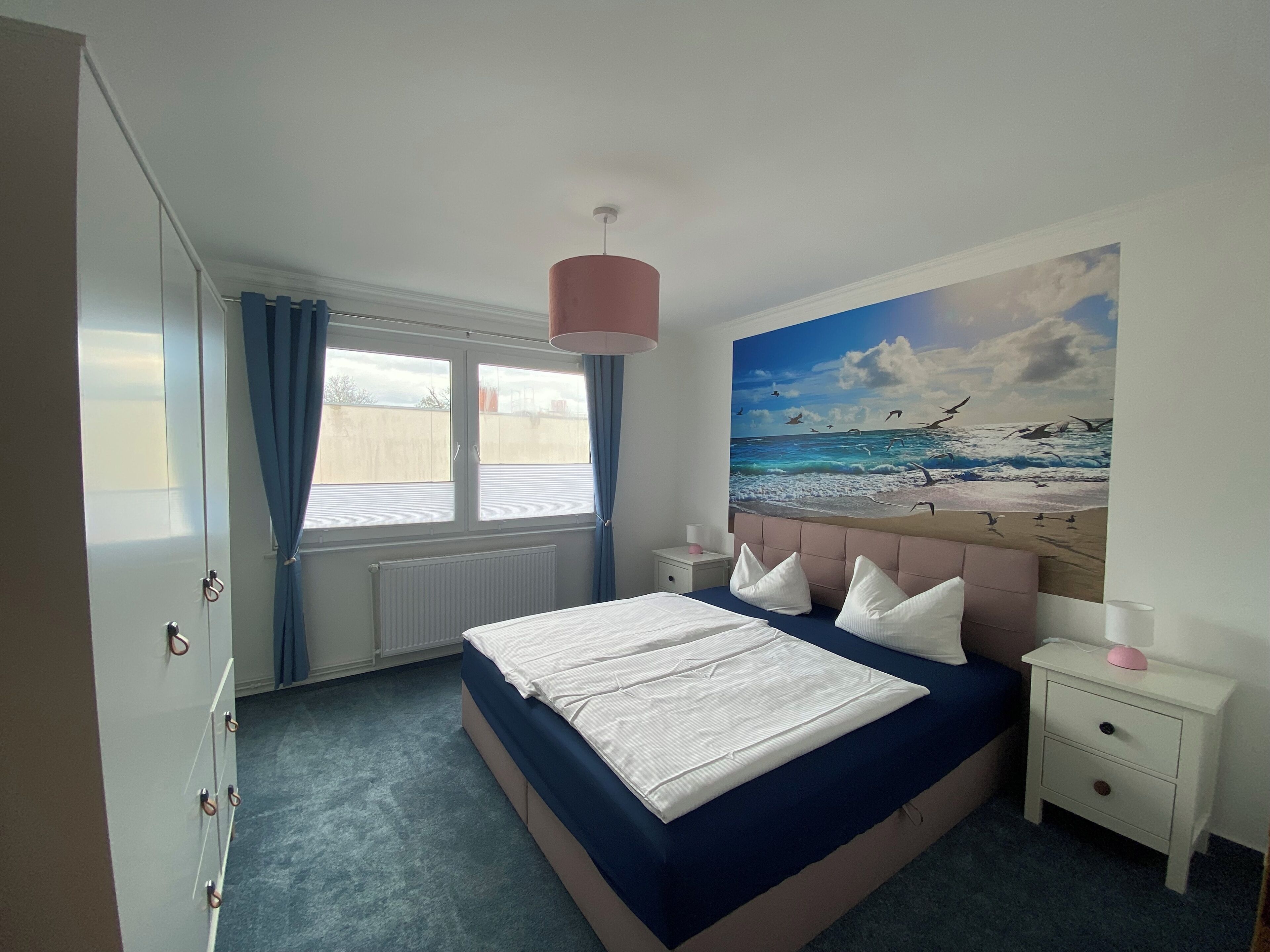 Un dormitorio con cama, mesita de noche, ventana con cortinas, radiador y un mural de playa en la pared.