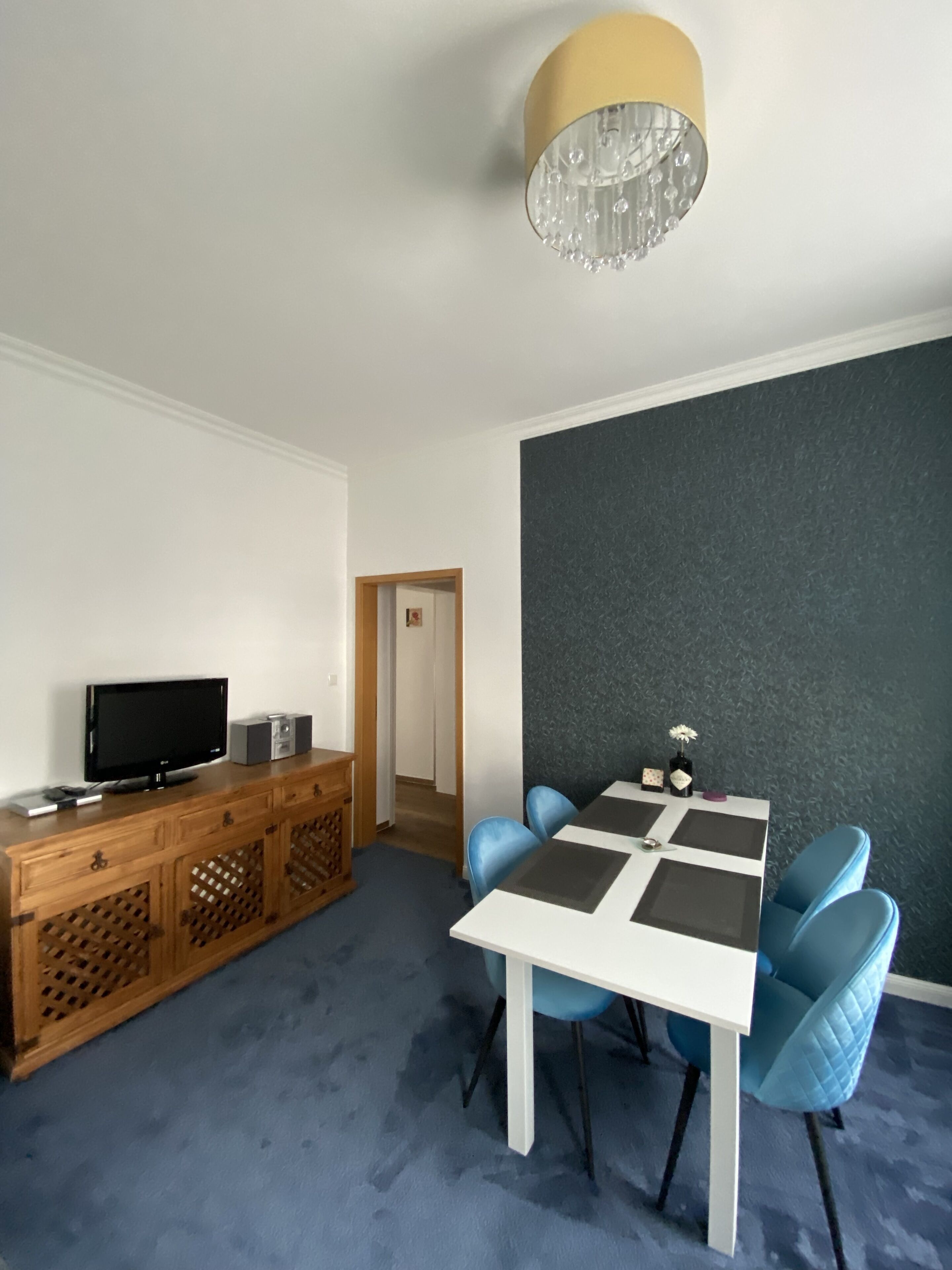 A modern living room with a wooden cabinet, a flat-screen TV, a table with blue chairs, a chandelier, and a dark textured wall.