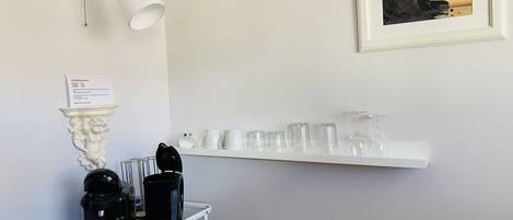 Une chambre d’hôtel avec un mur blanc, une machine à café fixée au mur, une étagère blanche avec des verres et un tableau représentant un paysage côtier.