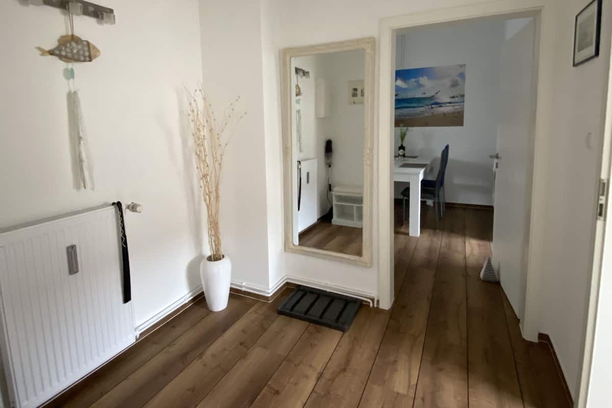 Un couloir doté d’un plancher en bois, d’un radiateur blanc, d’une plante en pot, d’un miroir et d’une porte donnant sur une pièce où se trouve un tableau.