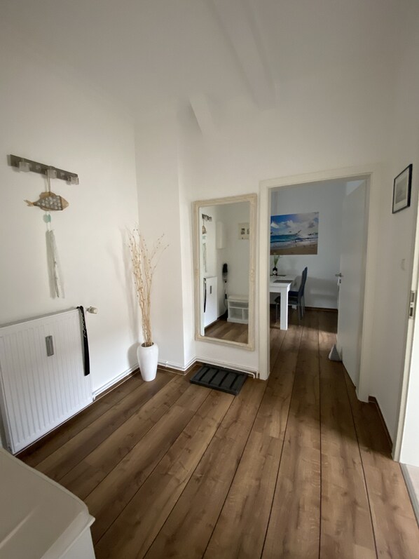 Un pasillo con suelo de madera, un radiador blanco, una planta en maceta, un espejo y una puerta que da a una habitación con un cuadro.