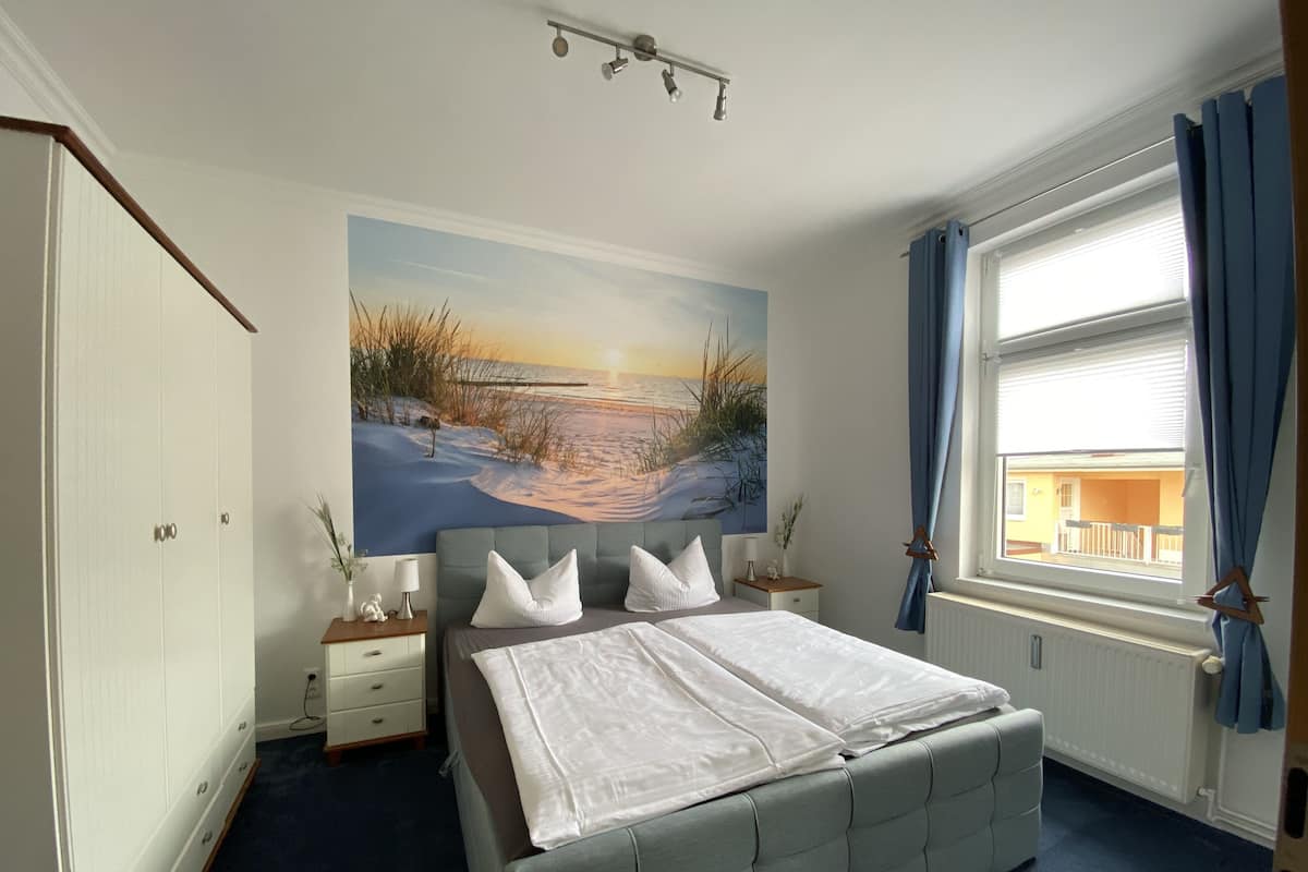 Une chambre à coucher avec un grand lit, une table de chevet, une commode, un tableau représentant une scène de plage et une fenêtre avec des rideaux.