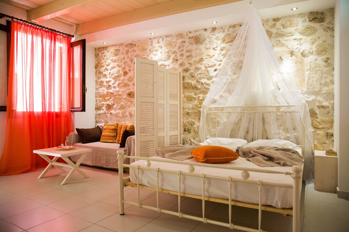Un dormitorio con cama, mesita de noche, una silla, una mesa y ventana con cortinas.