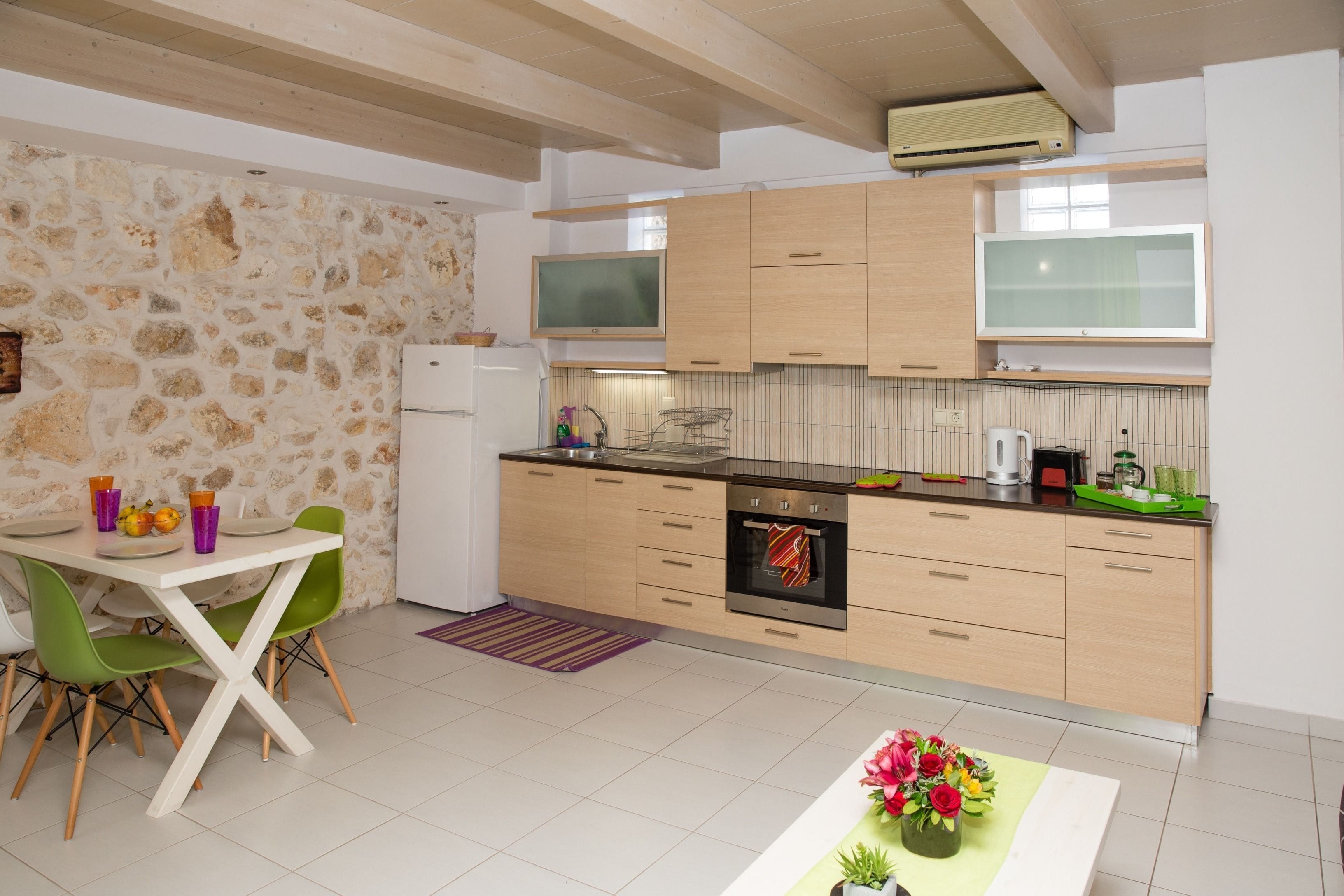 A modern kitchen with a stone wall, wooden cabinets, a built-in oven, and a dining area with green chairs and a table.