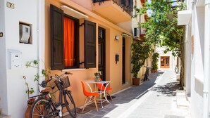 A narrow street with a bicycle parked by a white table and orange chairs, a potted plant, and a building with a balcony. - AC Homes Christos (Rethymno)