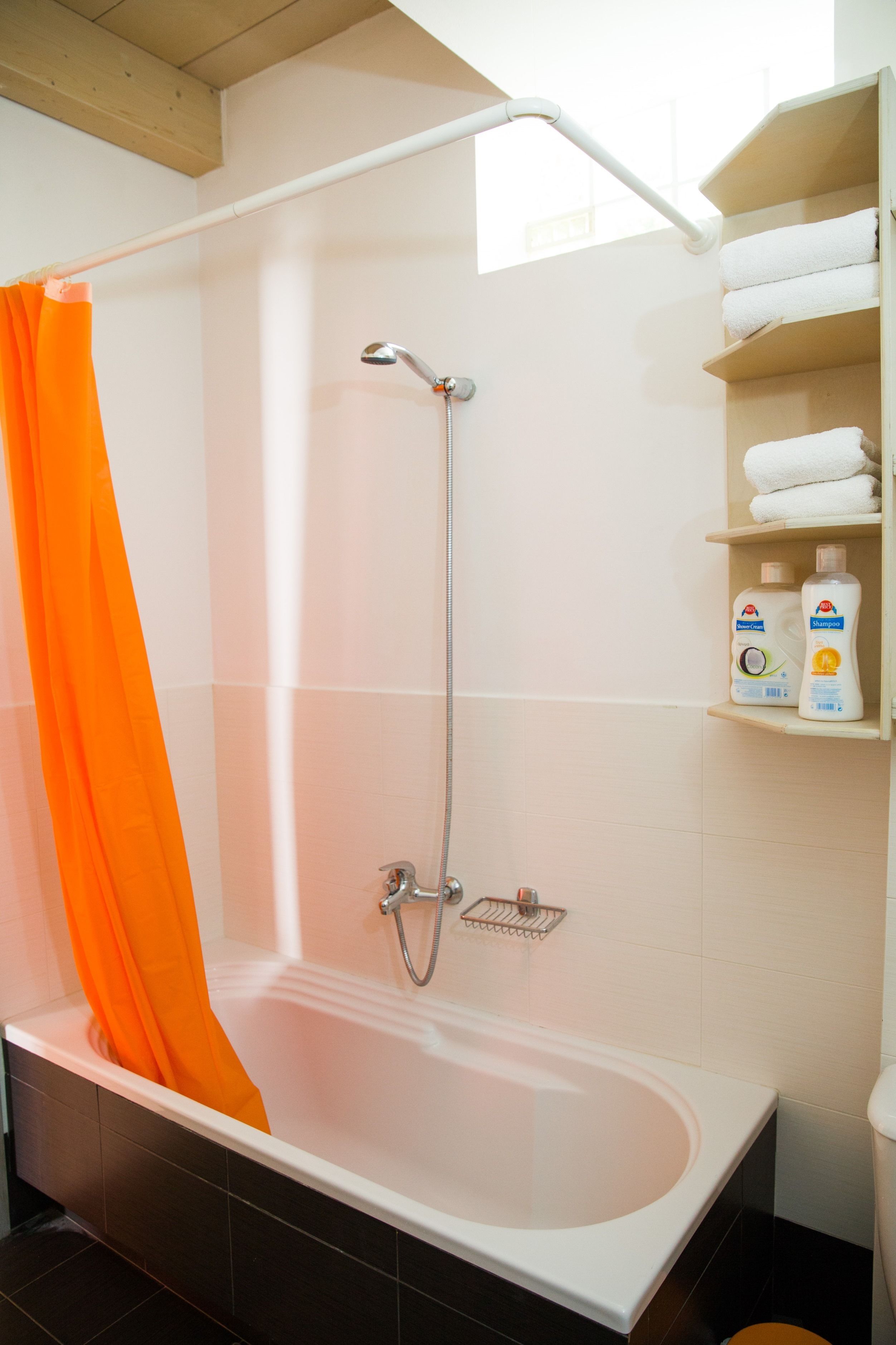 Un baño con bañera, una cortina de ducha naranja, un cabezal de ducha y repisas con artículos de aseo.