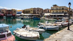A marina with boats docked, a waterfront promenade, and buildings with outdoor seating. - AC Homes Christos (Rethymno)