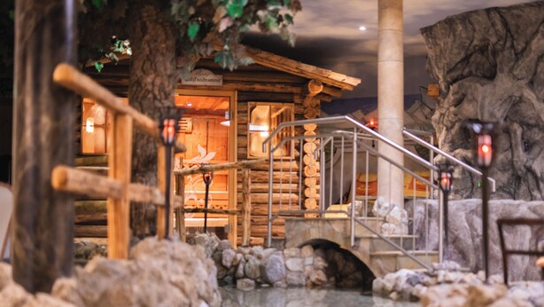 A rustic wooden cabin with a stone staircase leading to a wooden door, surrounded by rocks and a reflective water feature. - Landhotel Alpenhof (Filzmoos)