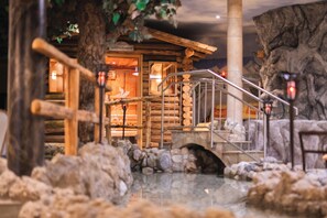 Una cabaña rústica de madera con una escalera de piedra que lleva a una puerta de madera, rodeada de rocas y un elemento acuático que refleja el paisaje.