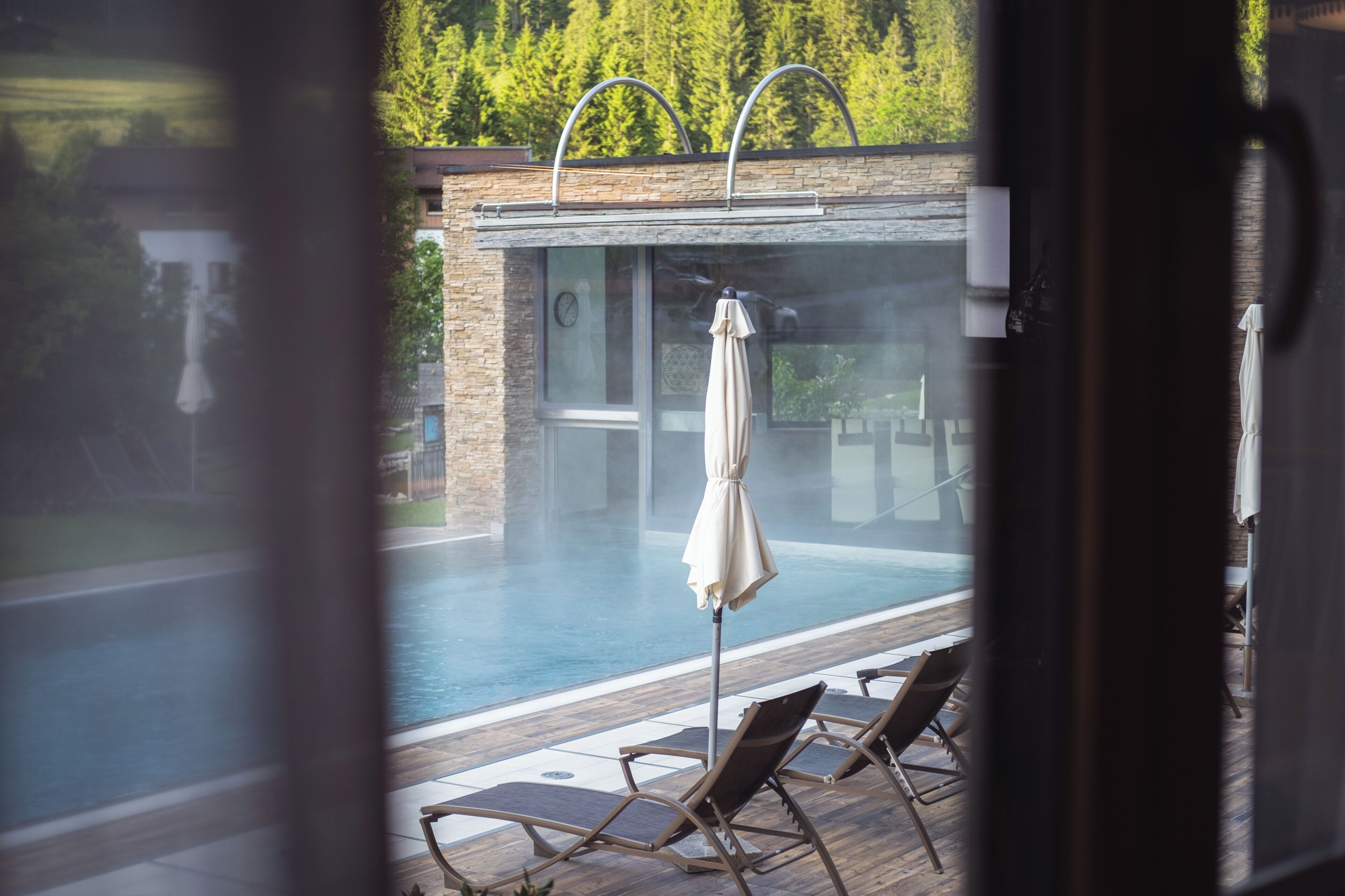 Une piscine avec des chaises longues et un parasol, entourée d’une terrasse en bois et d’un bâtiment en pierre.