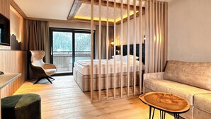 A modern hotel room with a bed, a sofa, a coffee table, a chair, and a view of the outdoors. - Landhotel Alpenhof (Filzmoos)