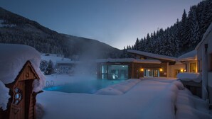 A snow-covered landscape with a heated pool, wooden cabins, and a mountain backdrop. - Landhotel Alpenhof (Filzmoos)
