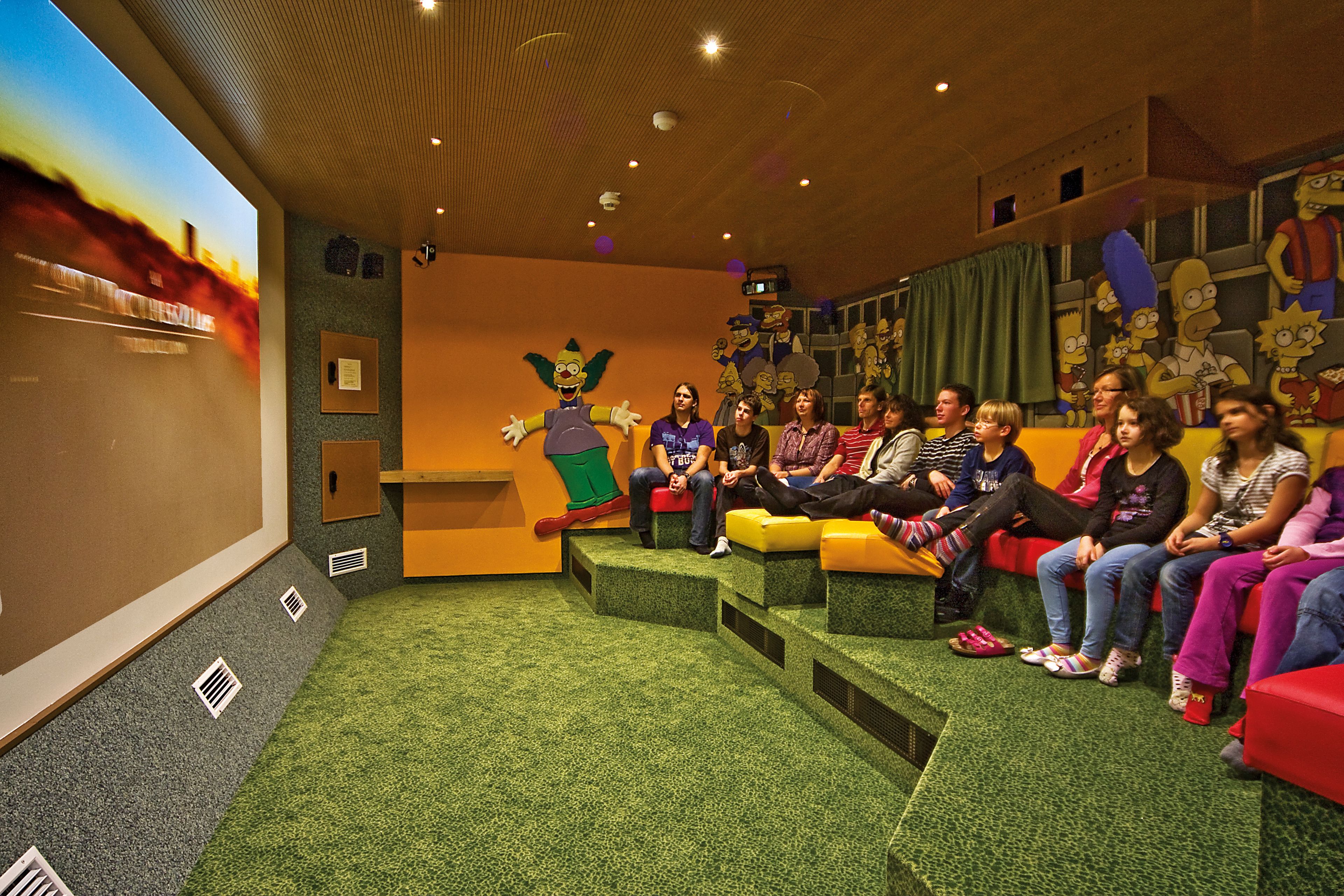 A group of people seated on colorful, low-rise steps in a room with a large mural of animated characters on the wall.