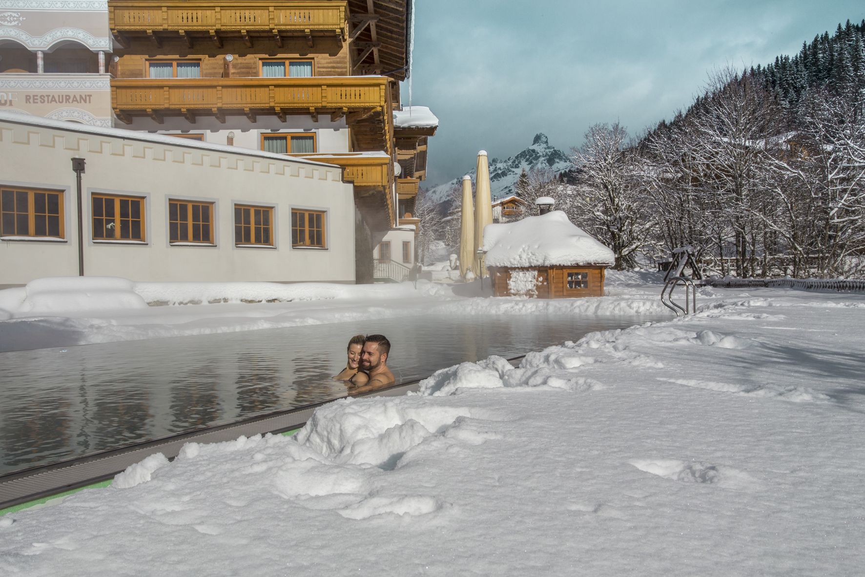 A snow-covered outdoor area with a pool, wooden buildings, and a mountain backdrop.