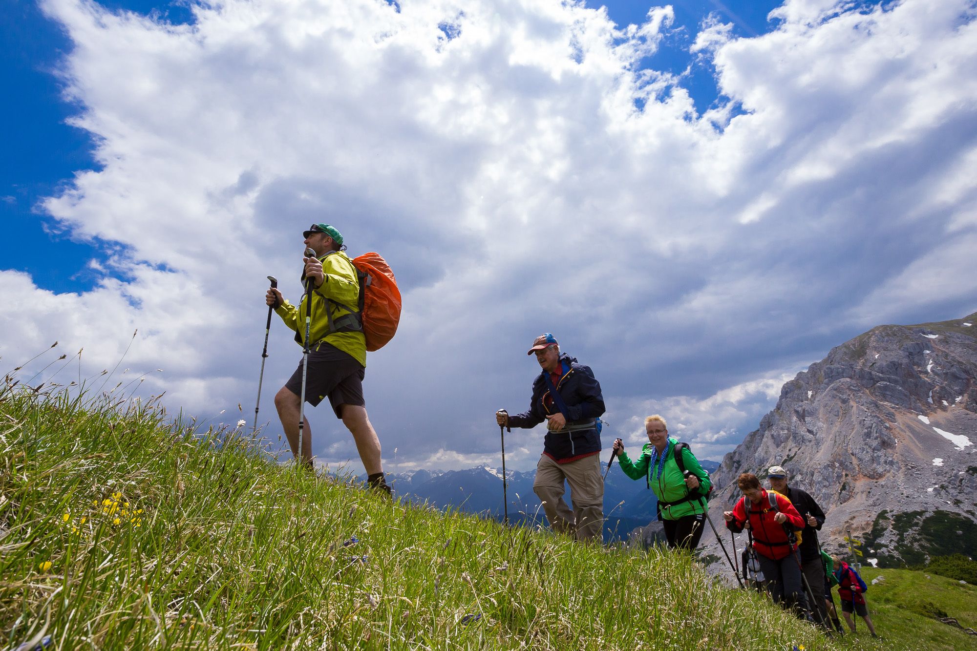A group of hikers on a grassy mountain trail with rocky terrain and mountains in the background.