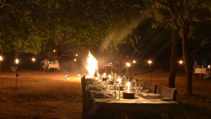 An outdoor dining setup with tables arranged in a semi-circle, surrounded by trees and illuminated by candles and lanterns.