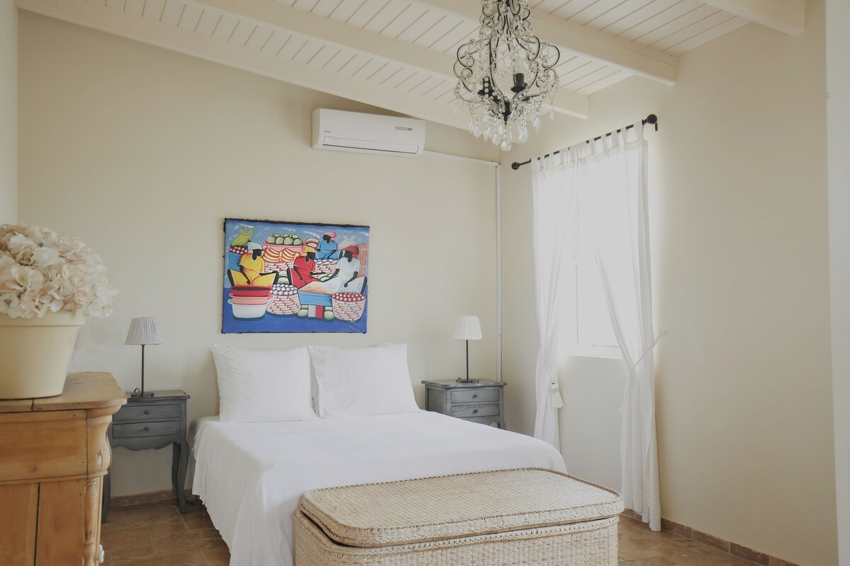 Un dormitorio con cama, mesita de noche, lámpara, un cuadro decorativo, una araña y una ventana con cortinas.