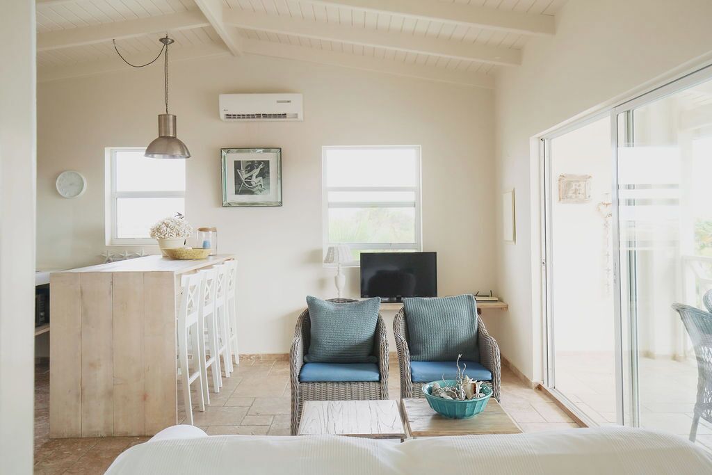 A bright, airy room with a wooden desk, two wicker chairs with cushions, a television, and a large window.