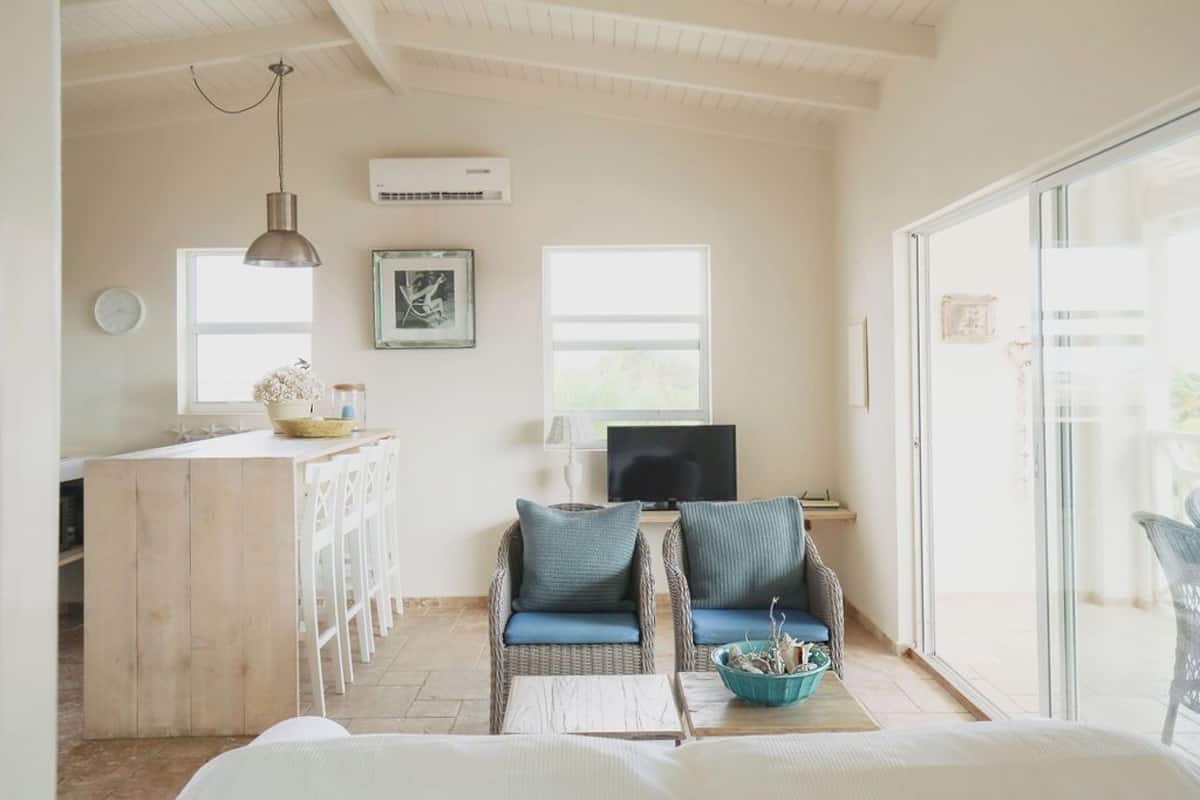 A bright, airy room with a wooden desk, two wicker chairs with cushions, a television, and a large window.