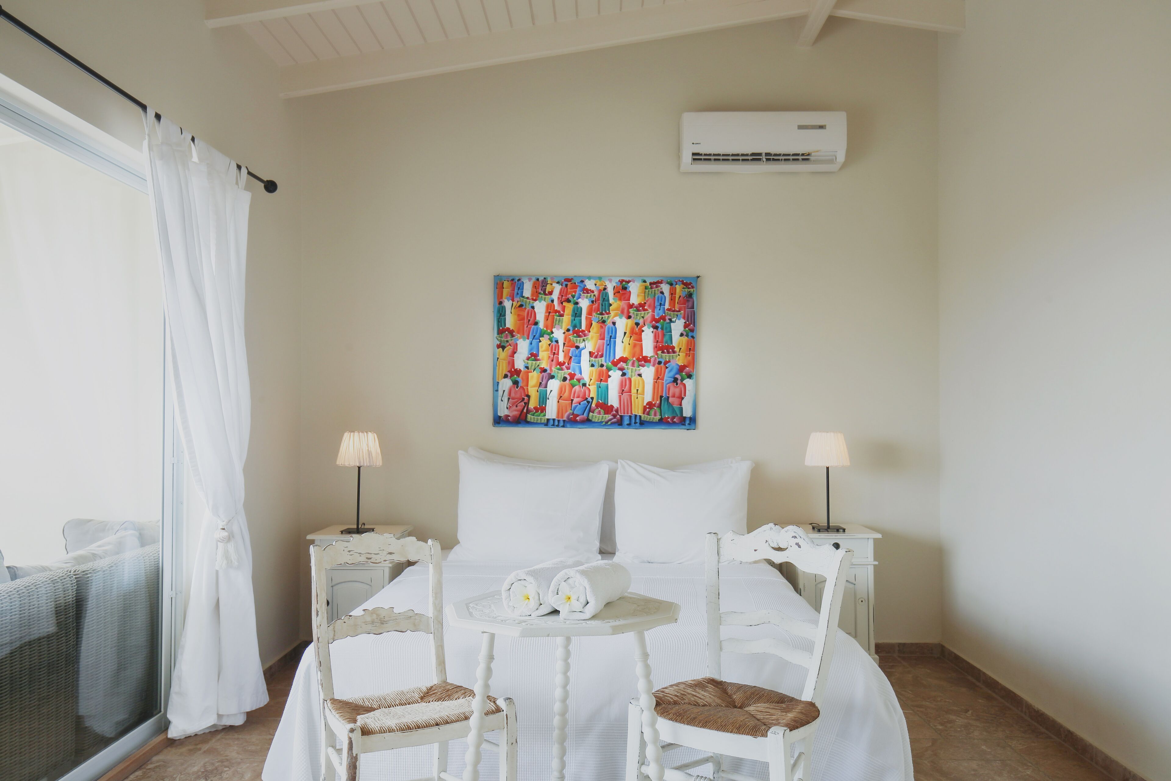 Une chambre d’hôtel comprenant un lit, deux tables de chevet, une petite table ronde, deux chaises, une œuvre d’art colorée accrochée au mur et un appareil de climatisation.