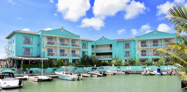 Un hôtel aux couleurs turquoise, doté de nombreux balcons, d’un espace piscine et de plusieurs bateaux amarrés.
