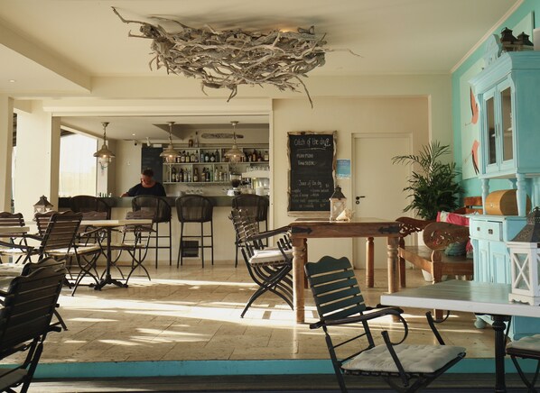 A restaurant with a bar area, wooden tables, and chairs. There is a chalkboard menu on the wall and a ceiling decoration made of branches. - Ocean Breeze Boutique Hotel & Marina (Kralendijk)