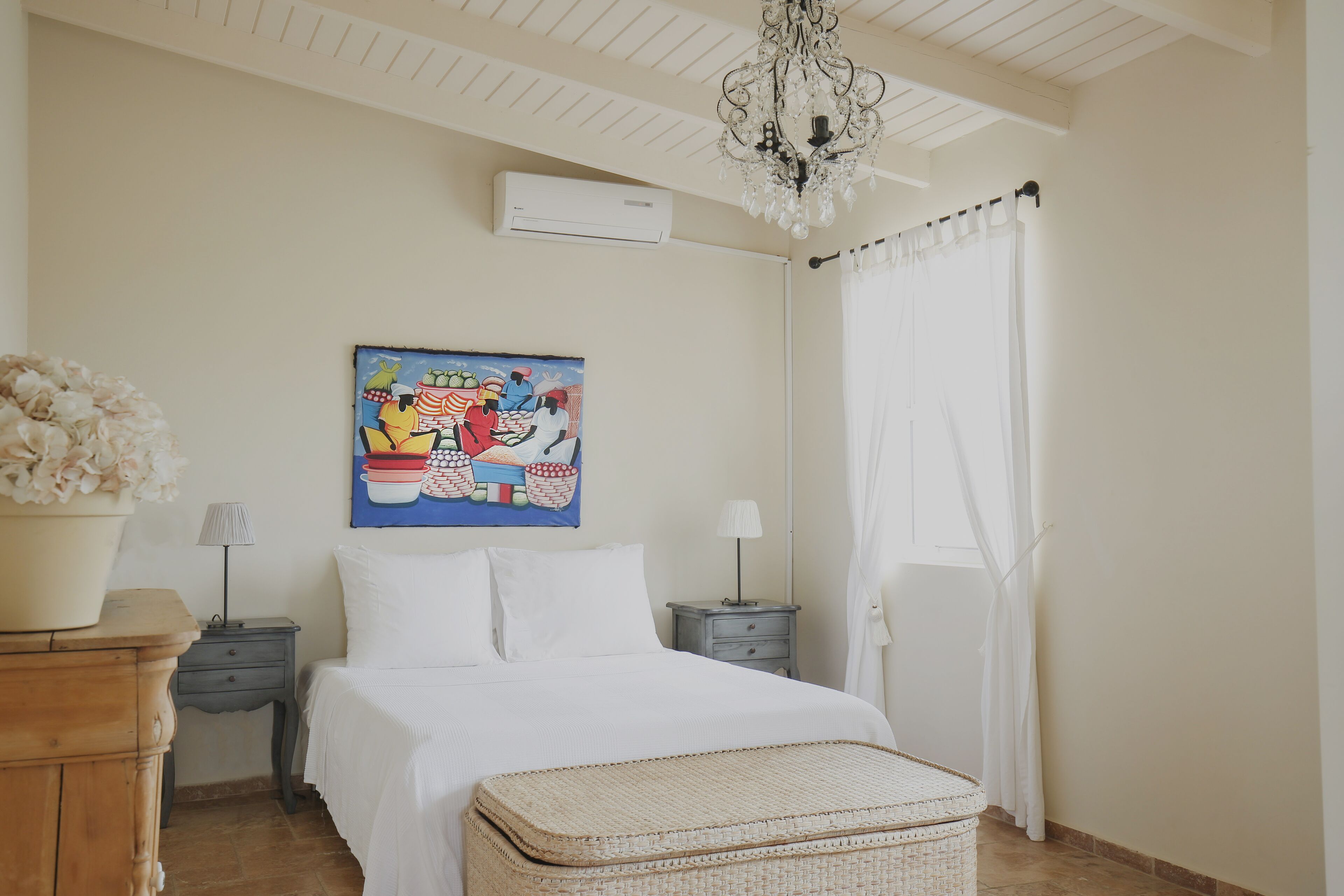 Een slaapkamer met een bed, een nachtkastje, een lamp, een decoratief schilderij, een kroonluchter en een raam met gordijnen.