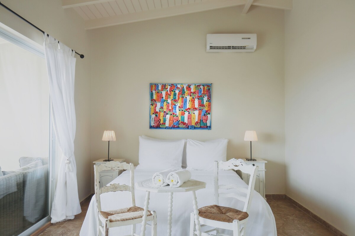 A hotel room with a bed, two bedside tables, a small round table, two chairs, a colorful artwork on the wall, and an air conditioning unit.