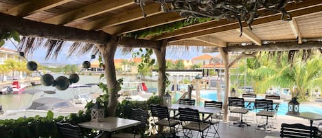 A covered outdoor seating area with tables and chairs, overlooking a pool and boats.