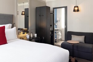 Une chambre d’hôtel comprenant un lit, un bureau avec une tasse et une bouilloire, une armoire, un canapé et une petite table ronde.