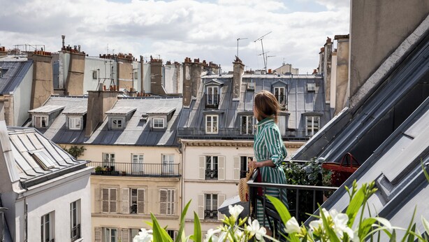 A balcony with a view of Parisian buildings, a woman standing, and some flowers in the foreground.