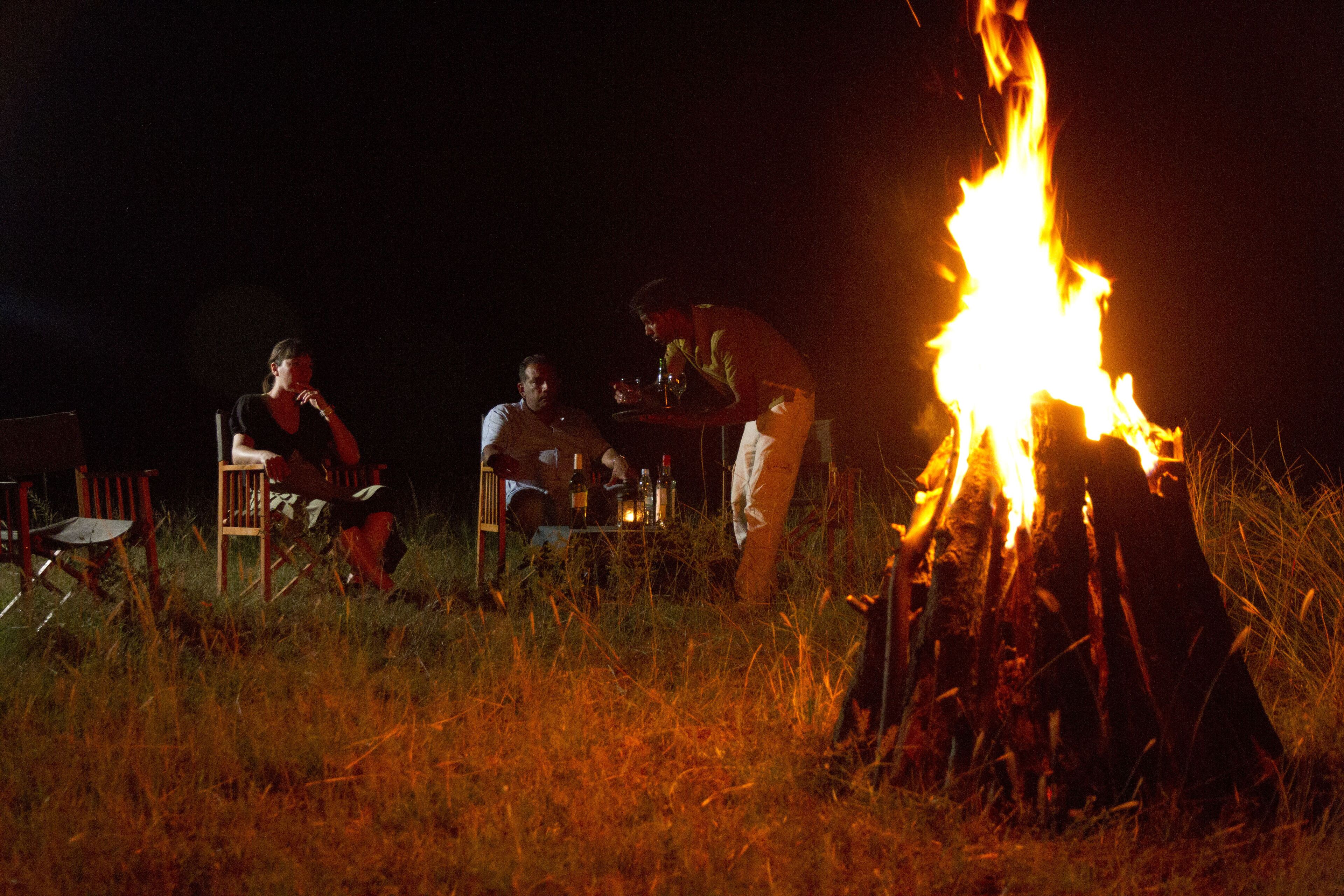 Un feu de camp autour duquel sont assis trois personnes, avec des chaises et des bouteilles, dans un espace herbeux, la nuit.