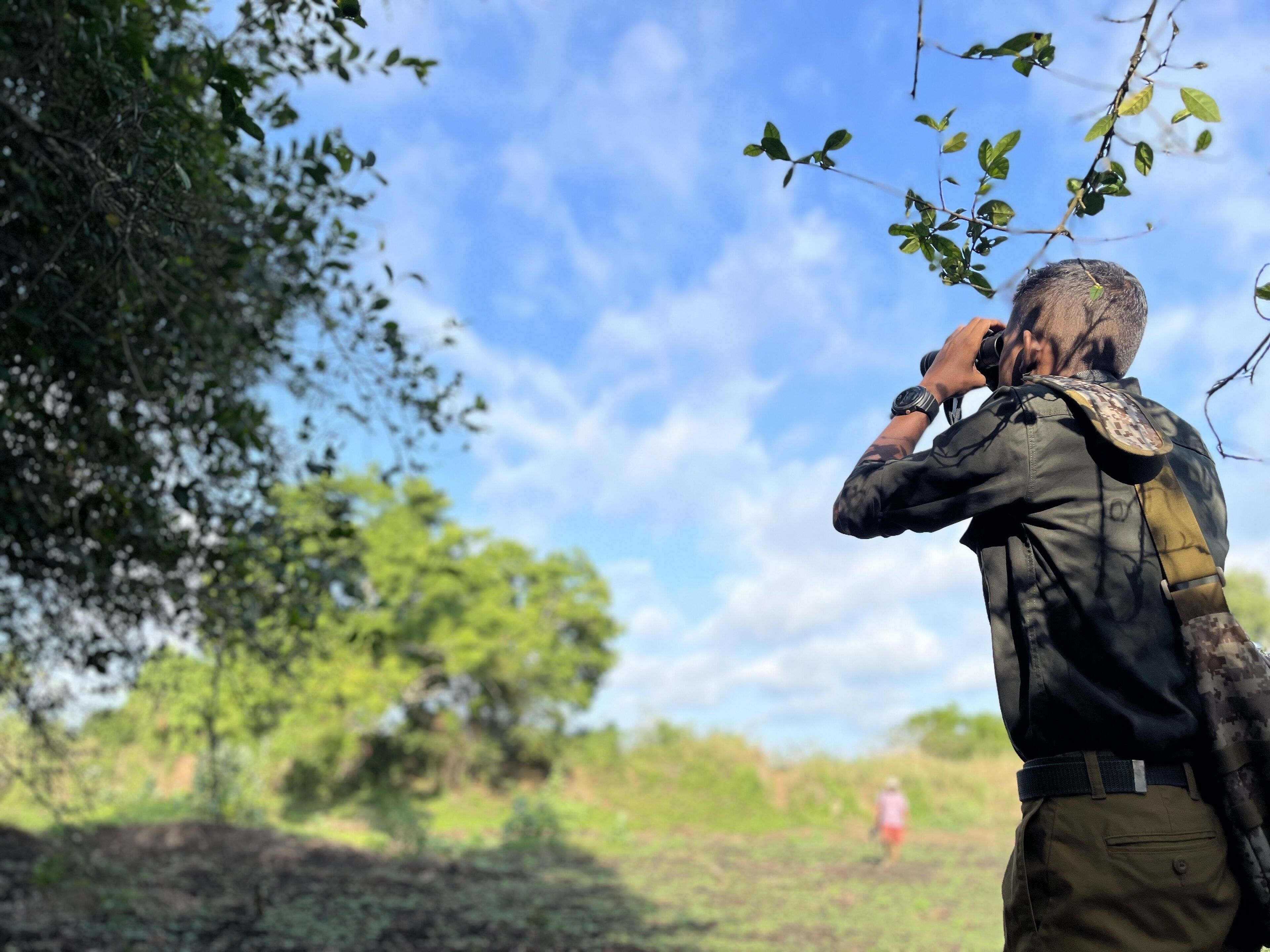 Una persona con uniforme militar, chaleco táctico y chaqueta de camuflaje, de pie al aire libre con árboles y cielo despejado de fondo.