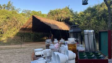 Un coin repas en plein air au style rustique, avec une cabane à toit de chaume, une table sur laquelle sont disposés de la vaisselle blanche, des tasses et un thermos, et une lanterne suspendue au-dessus.