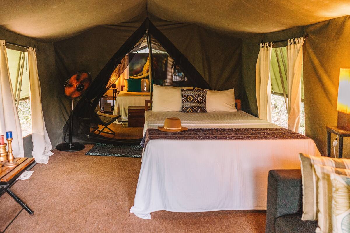 A cozy tented accommodation with a single bed, a small table with candles, and a fan.