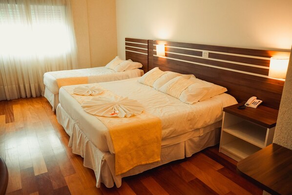 A hotel room with two beds, wooden flooring, a nightstand with a phone, and a window with sheer curtains.