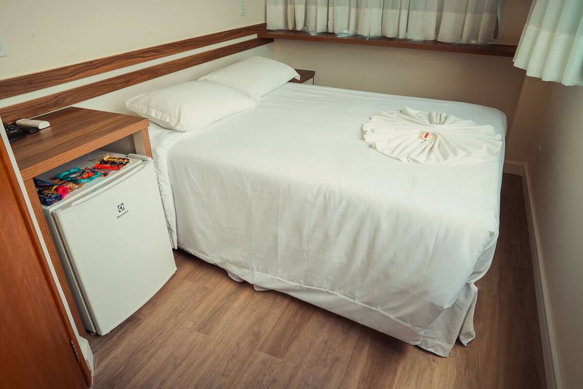 A neatly made bed with white linens, a small bedside table with a remote control and a power strip, and a compact refrigerator.