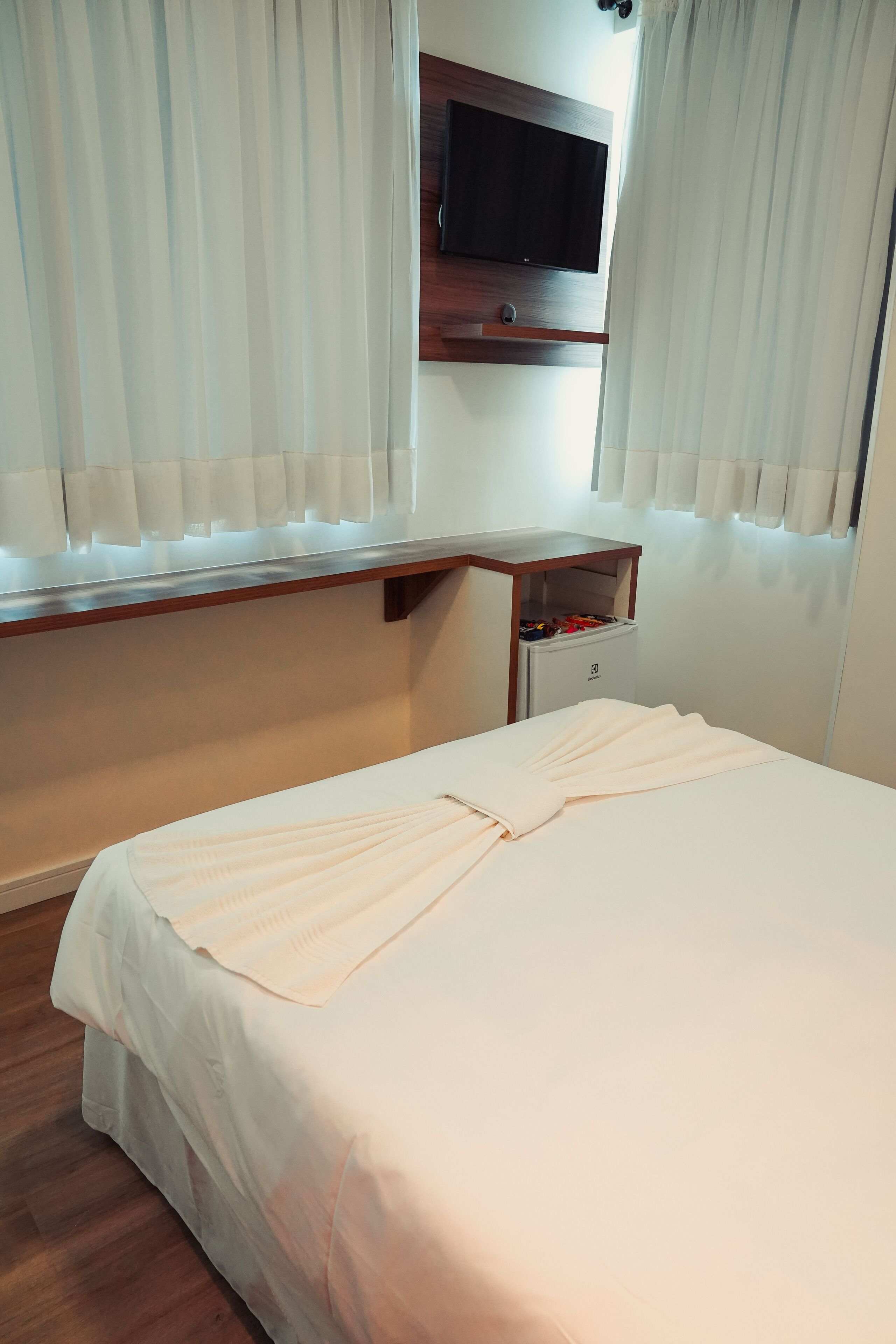 A hotel room with a bed, a TV mounted on the wall, a small desk with a chair, and a wardrobe.