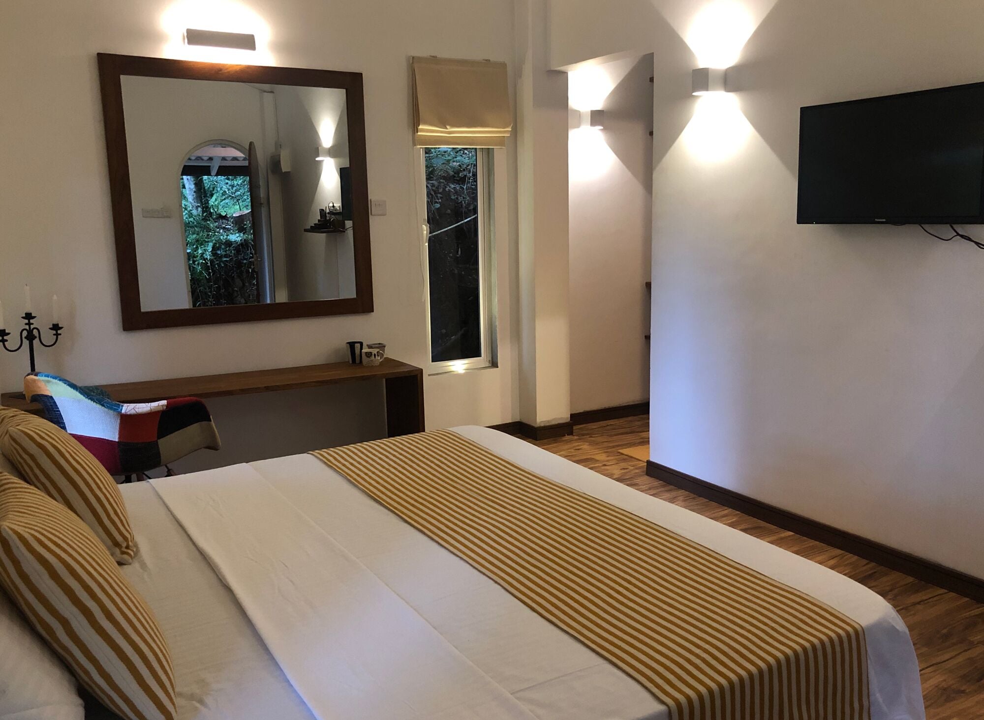 A hotel room with a large bed, a desk, a mirror, a television, and a window with a view of trees.