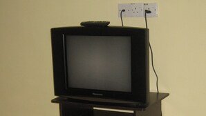 Un televisor sobre un soporte de madera, con un control remoto encima, conectado a un enchufe en la pared.