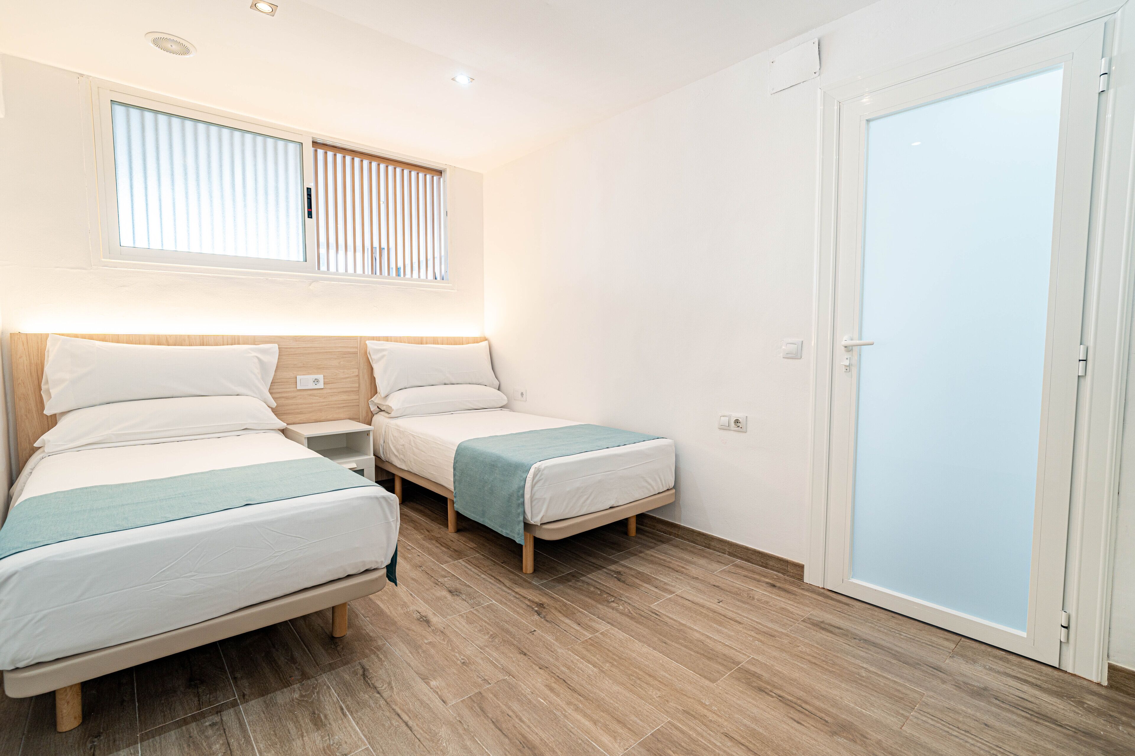 Habitación de hotel con dos camas, una ventana con persianas, suelo de madera y una puerta con vidrio esmerilado.