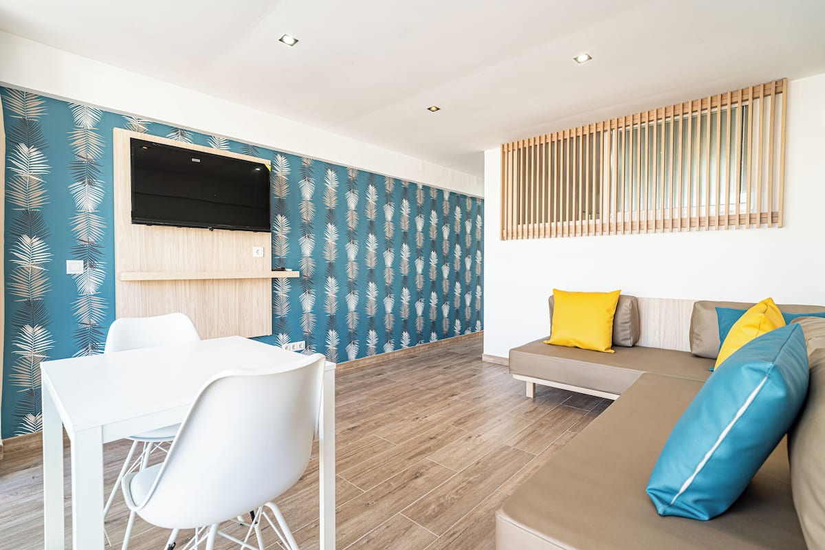 A modern living room with a white dining table, a sofa with blue and yellow cushions, and a patterned wall.