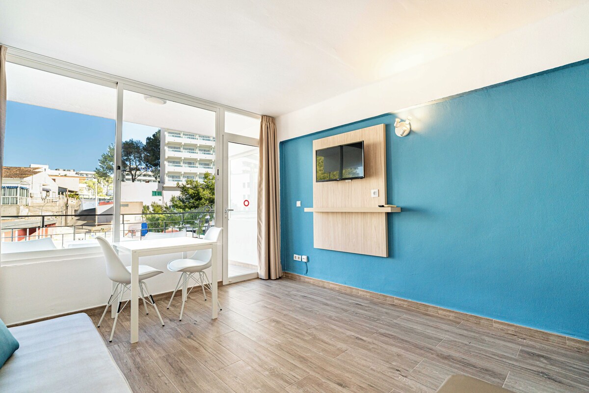 Een moderne kamer met een grote schuifglazen deur naar een buitenbalkon, een flatscreen televisie aan de wand en een klein houten plankje.