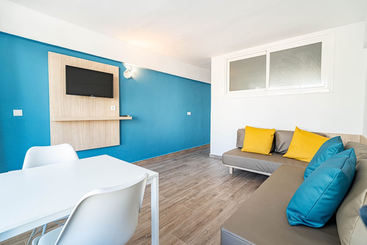 A modern living room with a blue accent wall, a flat-screen TV mounted on a wooden panel, a white dining table with chairs, and a sofa with yellow and blue cushions.