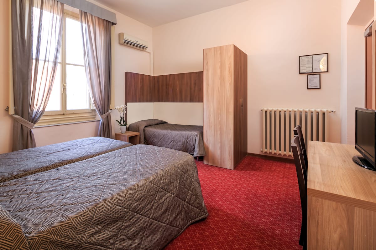 A hotel room with two beds, a desk, a TV, a radiator, and a window with curtains.