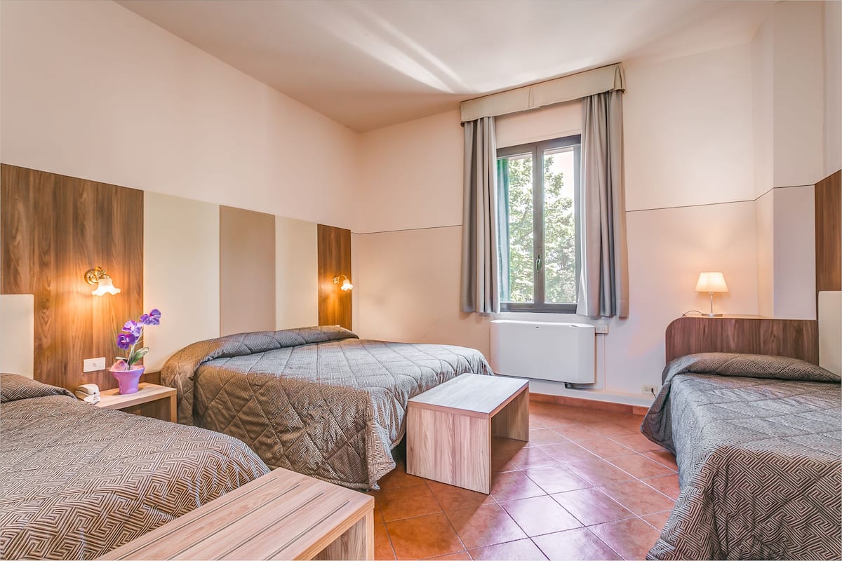 A hotel room with two beds, a small table, a nightstand with a lamp, and a window with curtains.