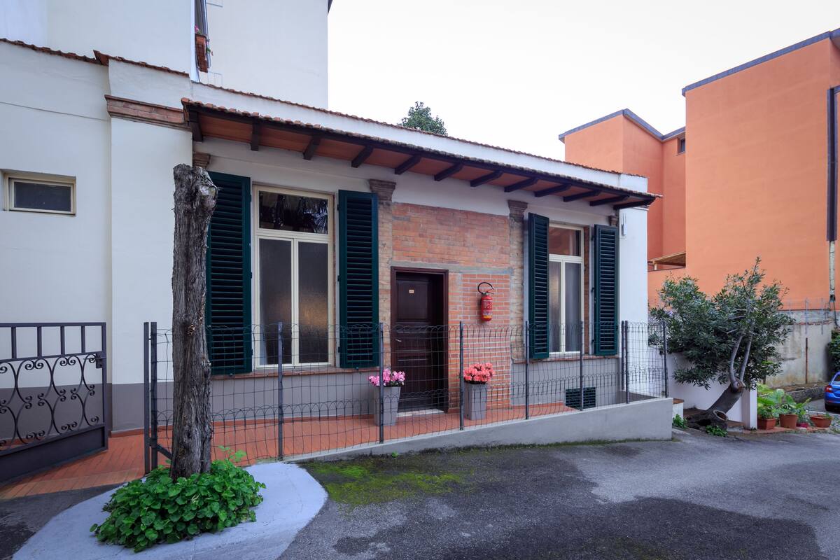 A two-story house with a brick facade, green shutters, and a small front garden with potted plants.
