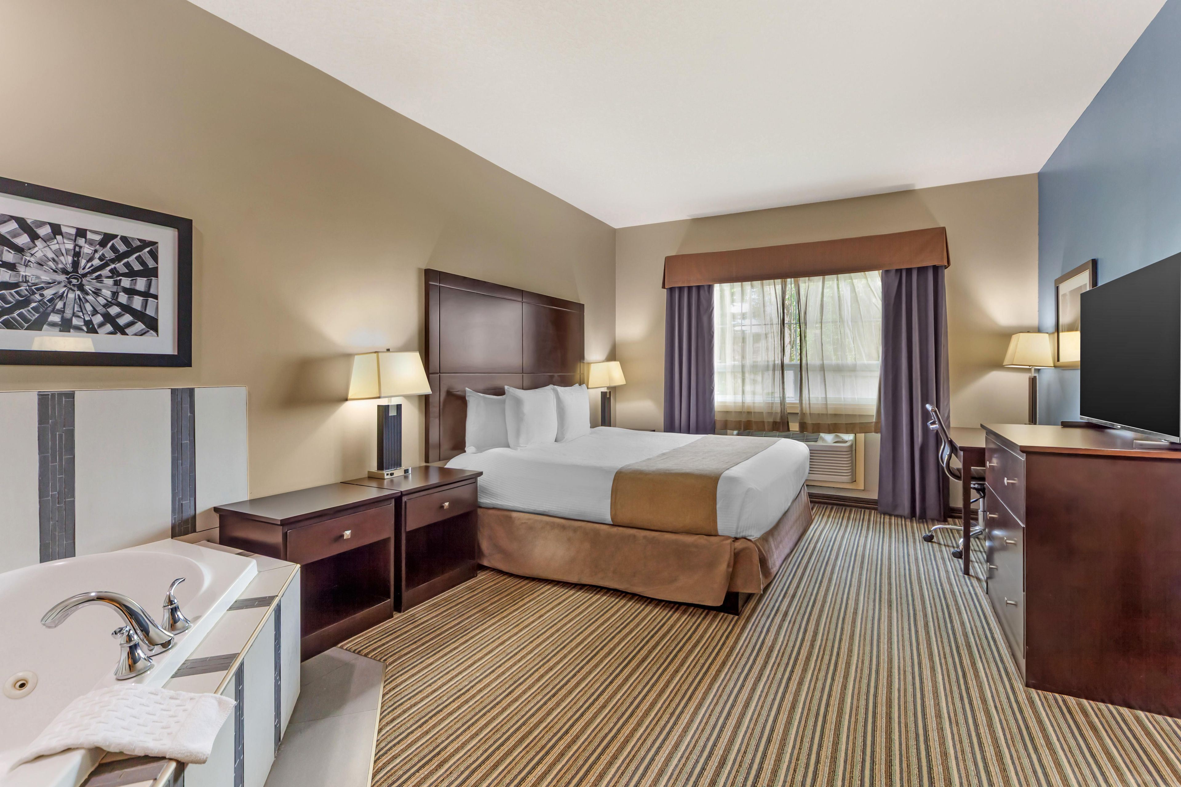 A hotel room with a large bed, two bedside tables, a desk with a chair, a flat-screen TV, a large window with curtains, and a bathroom with a sink and mirror.